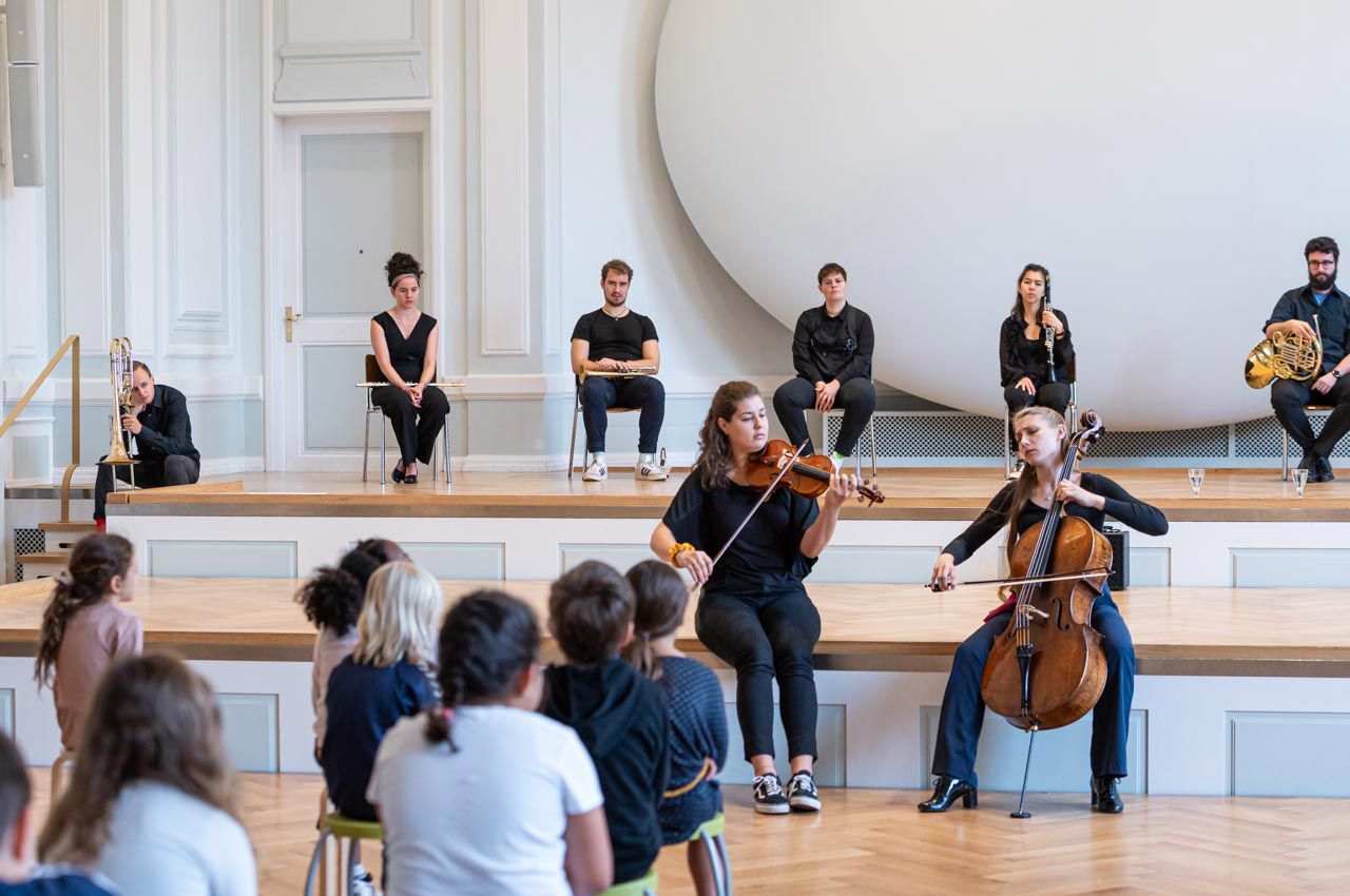 Zwei Studenten zeigen einem Mädchen die Tuba. Das Mädchen versucht zu spielen.